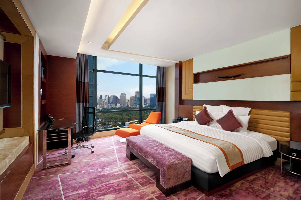 A modern hotel room with a large bed, a desk, a chair, and a view of the cityscape.