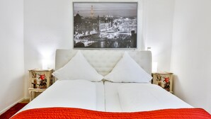 A neatly made bed with white linens and a red blanket, flanked by two bedside tables with decorative items, in a room with a cityscape view. - Hotel Laterne (Baden-Baden)