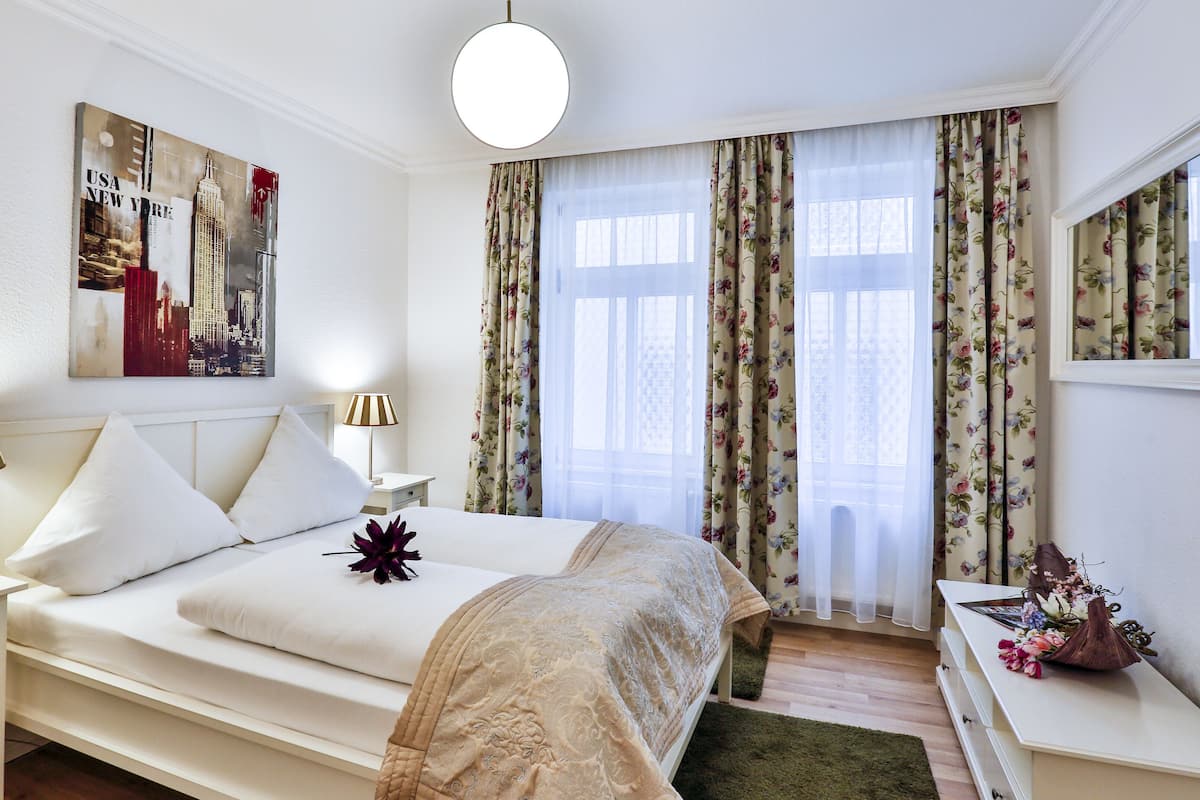 A hotel room with a neatly made bed, a bedside table with a lamp, a nightstand, and a floral curtain.