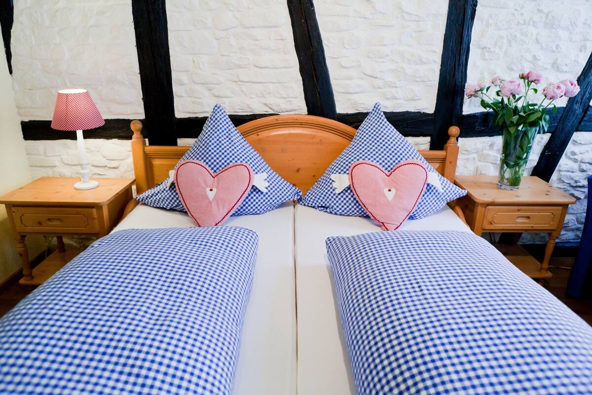 A double bed with checkered bedding, two pillows with heart designs, a bedside table with a lamp, and a vase with flowers.