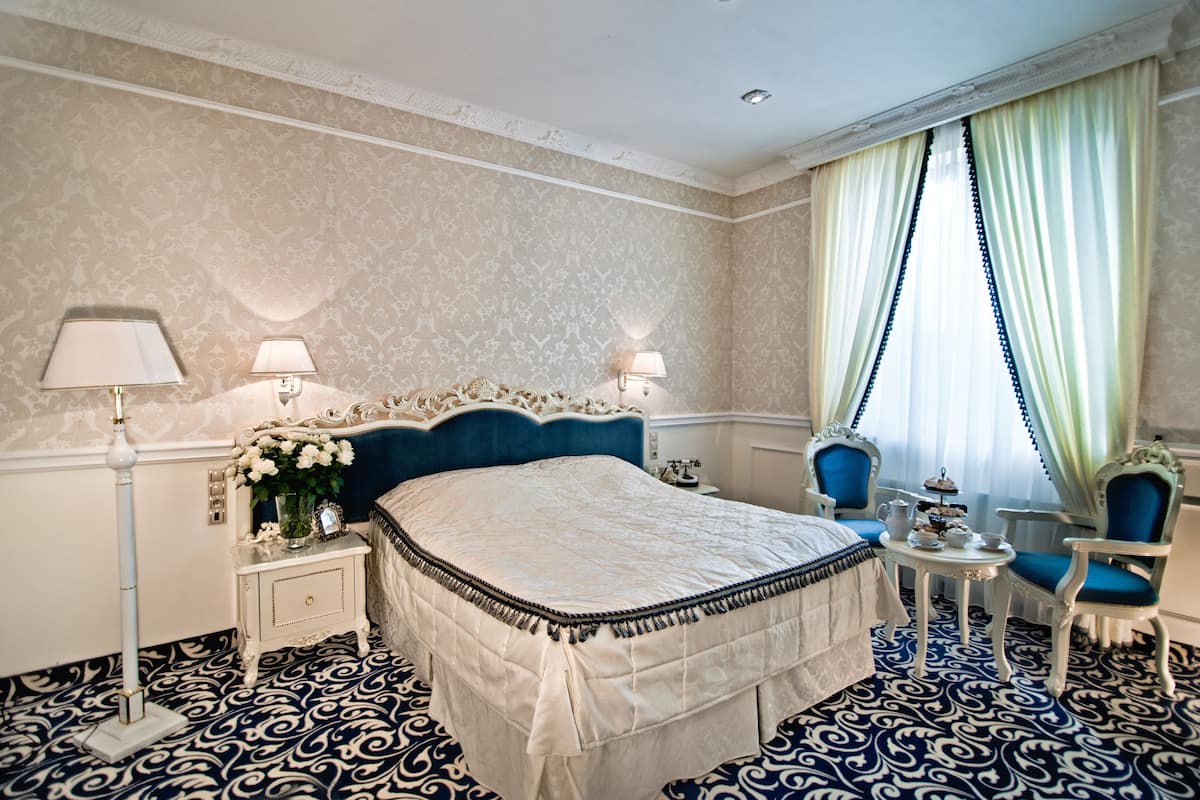 Una habitación de hotel con una cama grande, dos lámparas de noche, una mesita de noche con un jarrón de flores, una pequeña mesa con una tetera y tazas, y una silla.