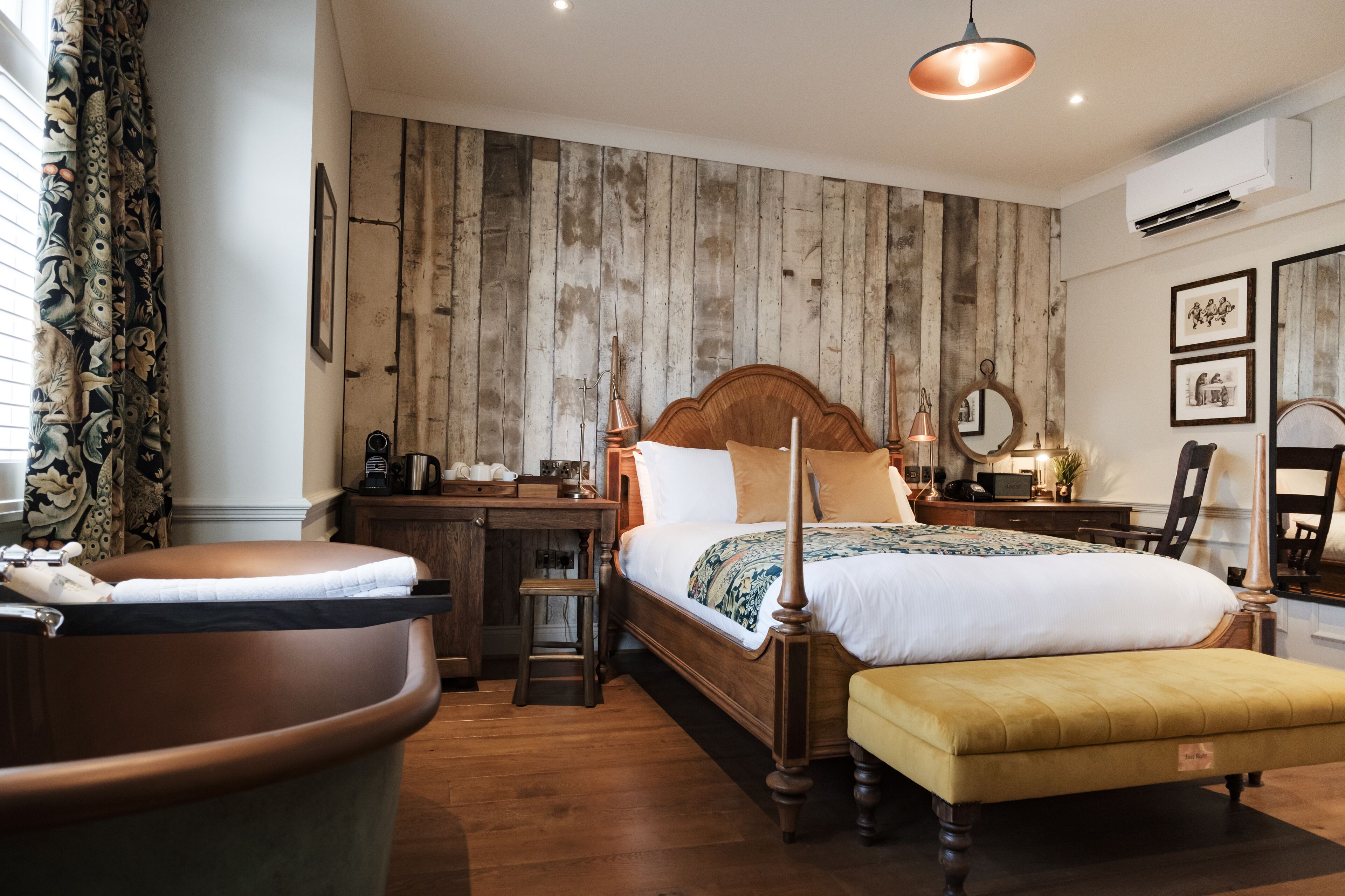 A bedroom with a wooden headboard, a four-poster bed, a bedside table, a nightstand, a large window with curtains, and a wooden floor.