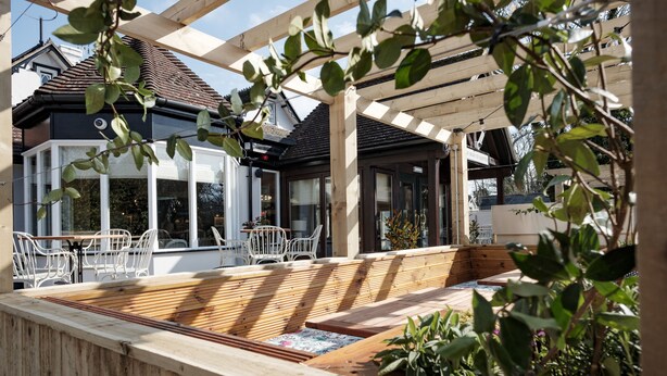 Um deck de madeira com área de estar integrada, rodeado por plantas em vasos e uma estrutura de pérgola.