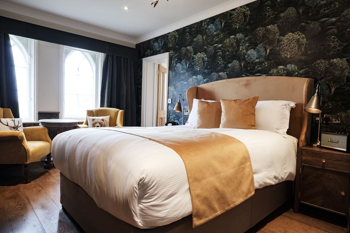 A hotel room with a large bed, a small round table, a chair, and a nightstand with a lamp.