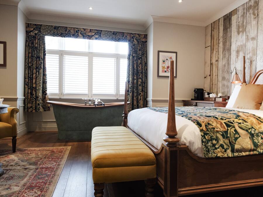 A bedroom with a large bed, a wooden headboard, a sitting area with a chair and table, a bathtub, and a window with curtains.