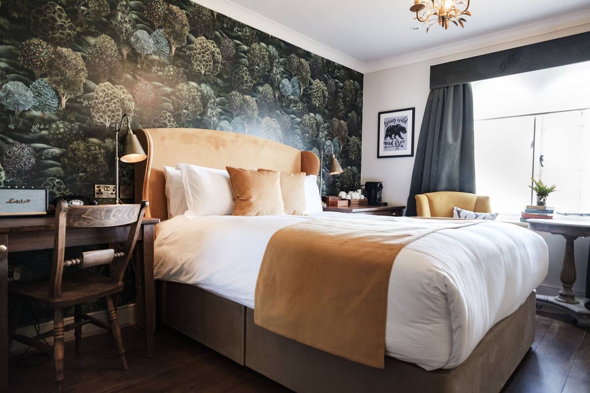 A hotel room with a large bed, a desk with a chair, a desk lamp, a window with curtains, and a wall with a nature-themed mural.