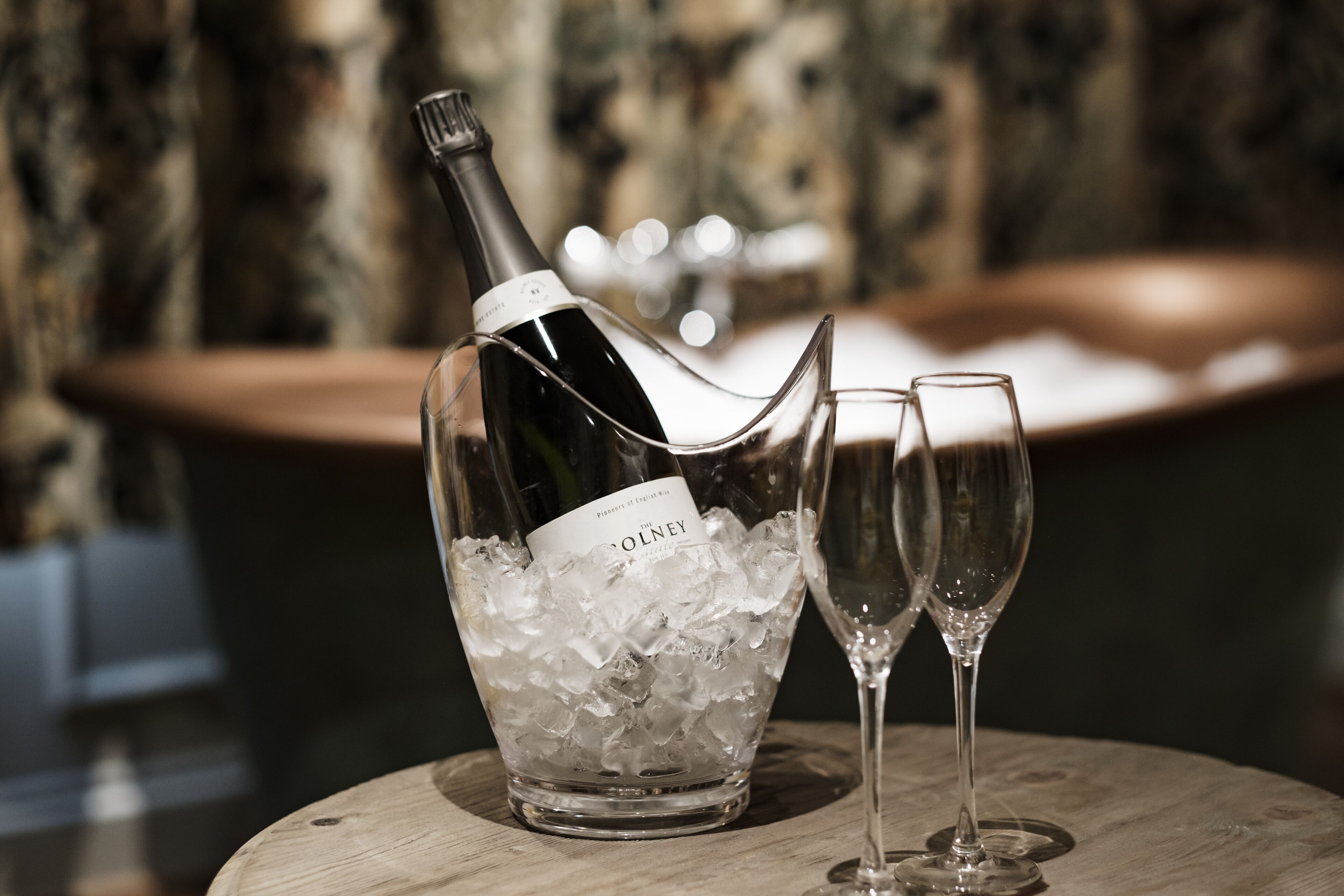 A bottle of wine in a glass container filled with ice cubes, with two empty wine glasses beside it.