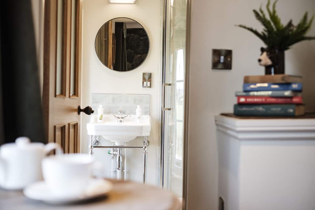 A bathroom with a white sink, a round mirror, and a glass shower door. There are books stacked on a small white cabinet to the right.