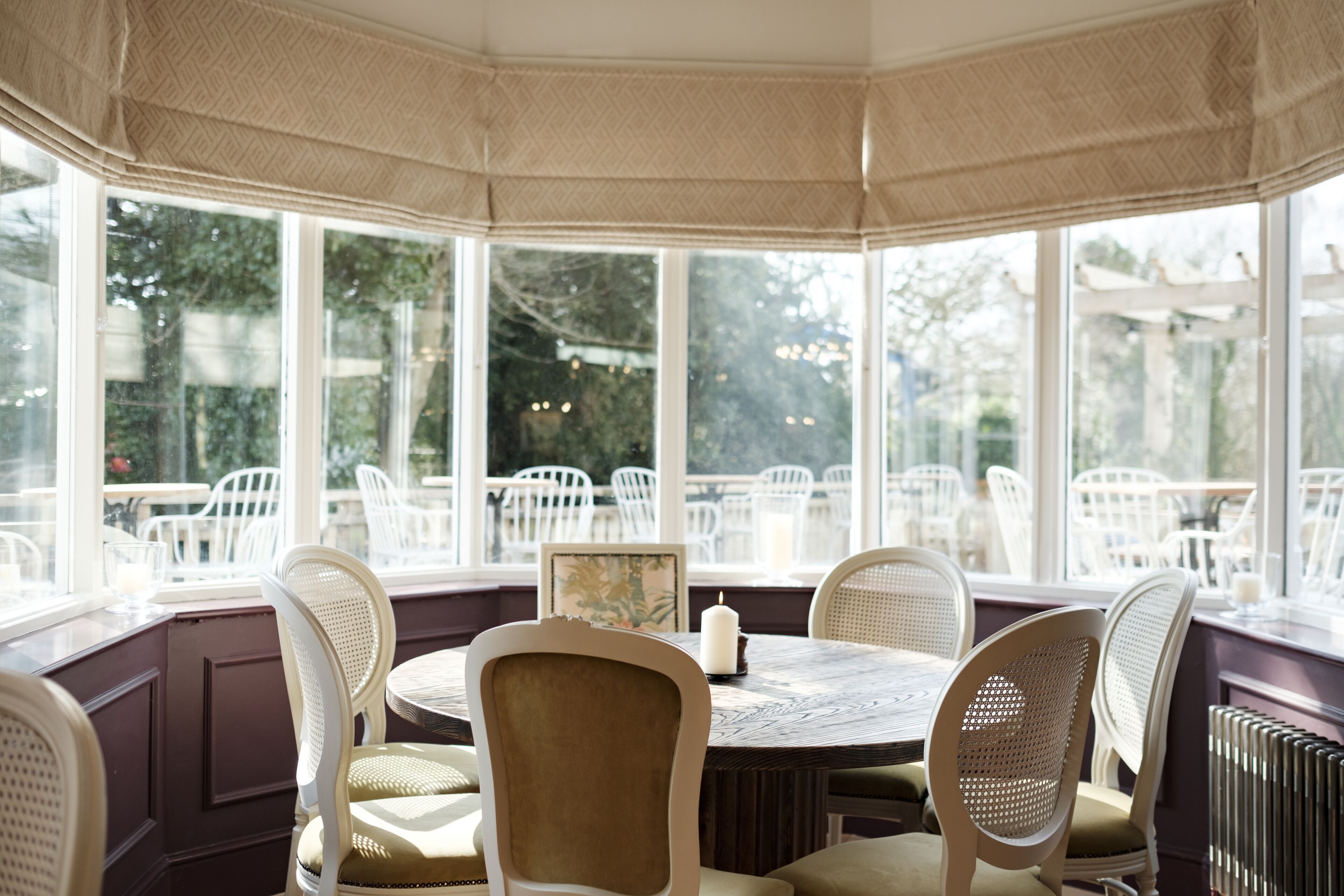 A bay window seating area with white wicker chairs and a round table, overlooking a garden with outdoor seating.