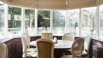 A bay window seating area with white wicker chairs and a round table, overlooking a garden with outdoor seating.