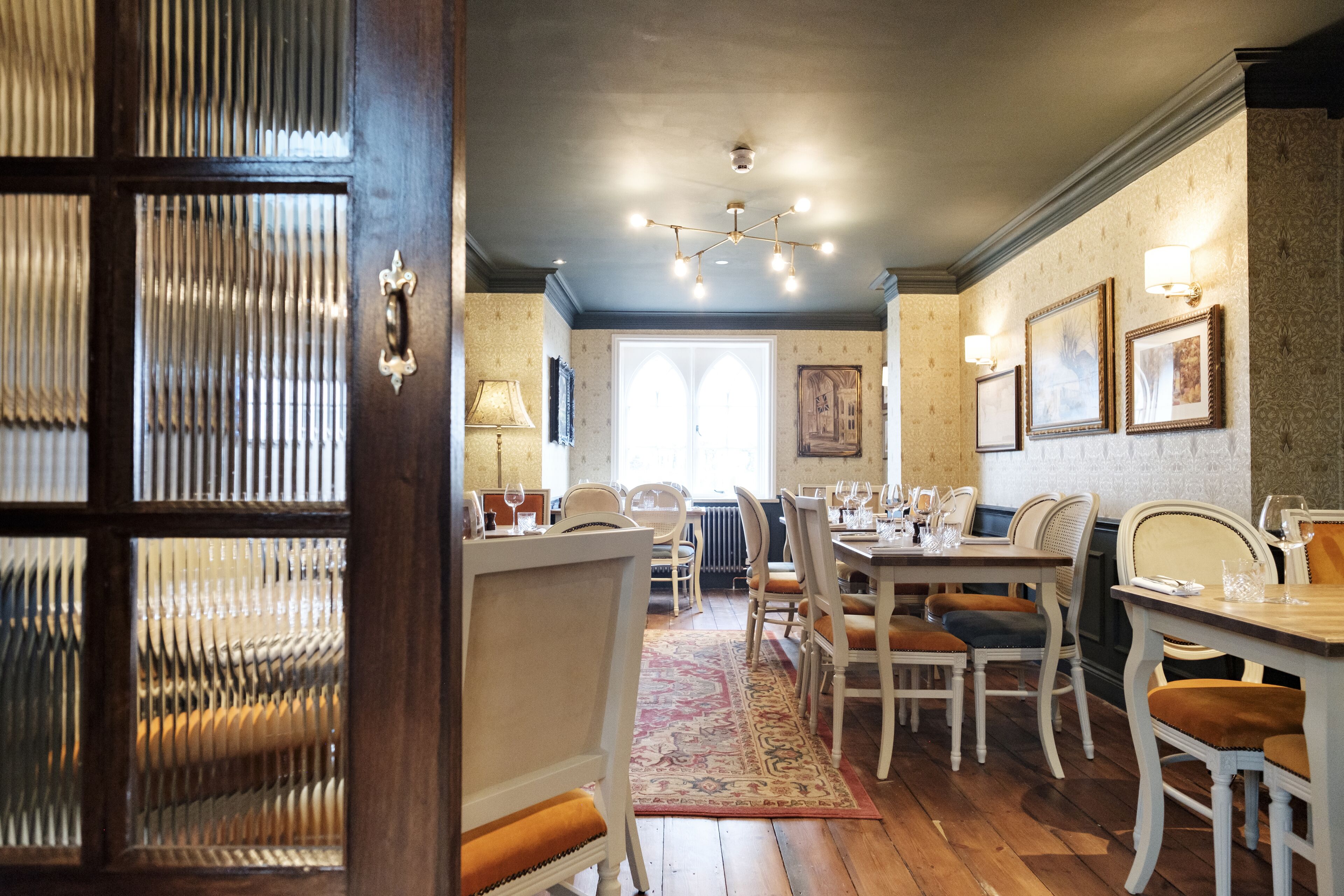 A dining area with wooden tables and chairs, a patterned rug, and wall-mounted light fixtures.