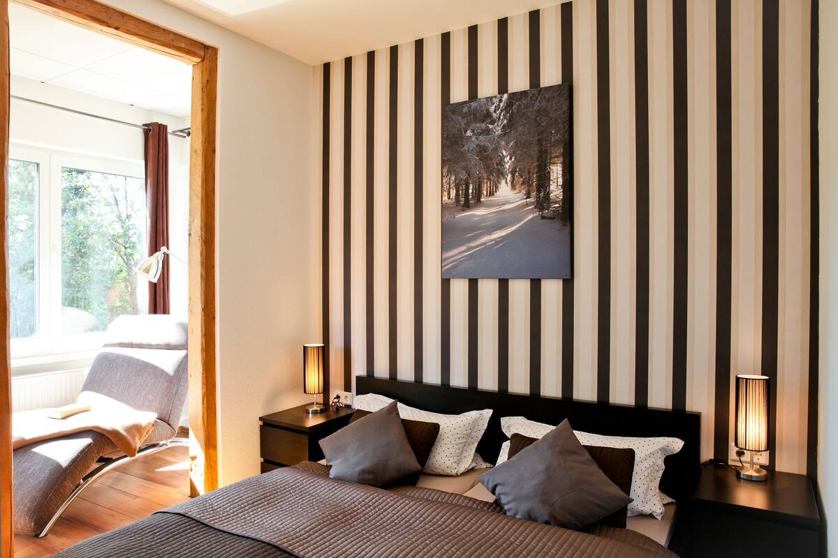 Un dormitorio con una cama, almohadas, una mesita de noche, una lámpara, una silla y una ventana.