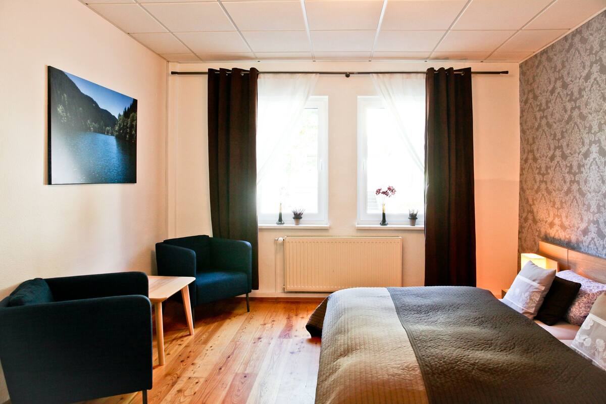 Una habitación de hotel con una cama, dos sillas, una mesita y una ventana con cortinas.