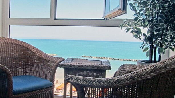 Una silla de mimbre y una mesa junto a un ventanal con vistas a una playa y un rompeolas rocoso.