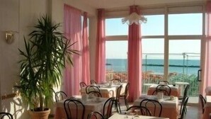 Un comedor con mesas dispuestas para dos, ventanas amplias con cortinas y una planta en maceta.