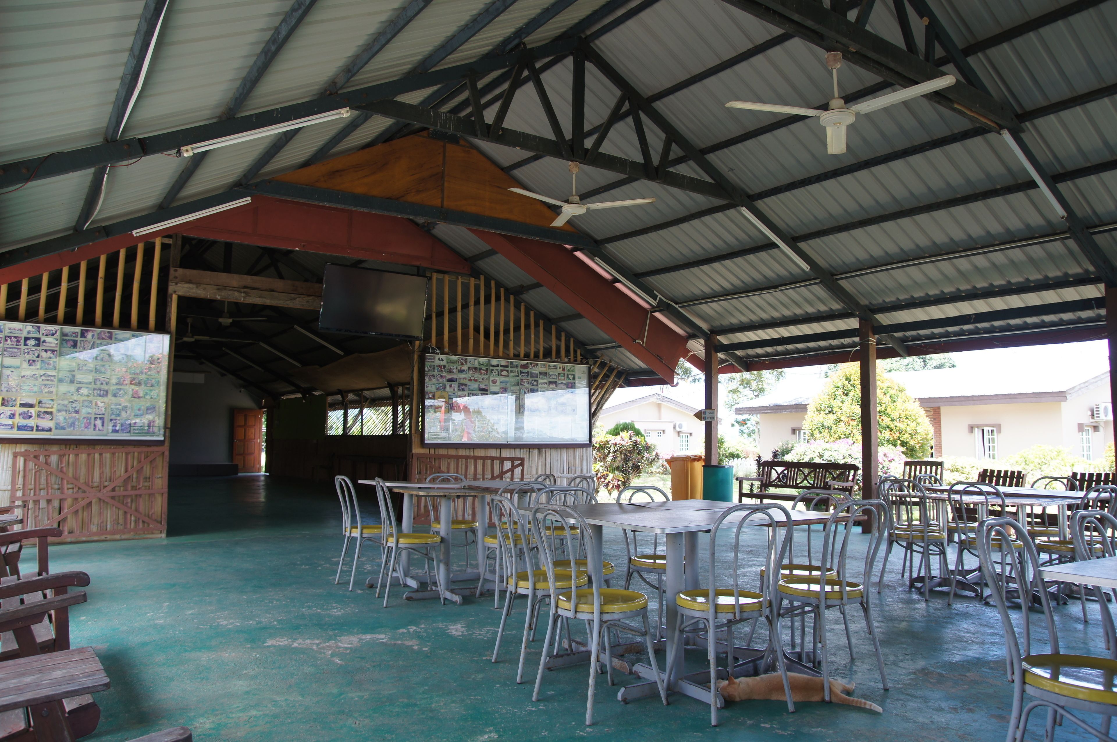 An indoor dining area with metal tables and chairs, a ceiling fan, and a bulletin board with various notices.