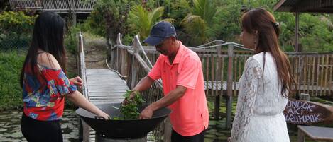 Tres personas cerca de una olla grande al aire libre, con un puente de madera y vegetación de fondo.