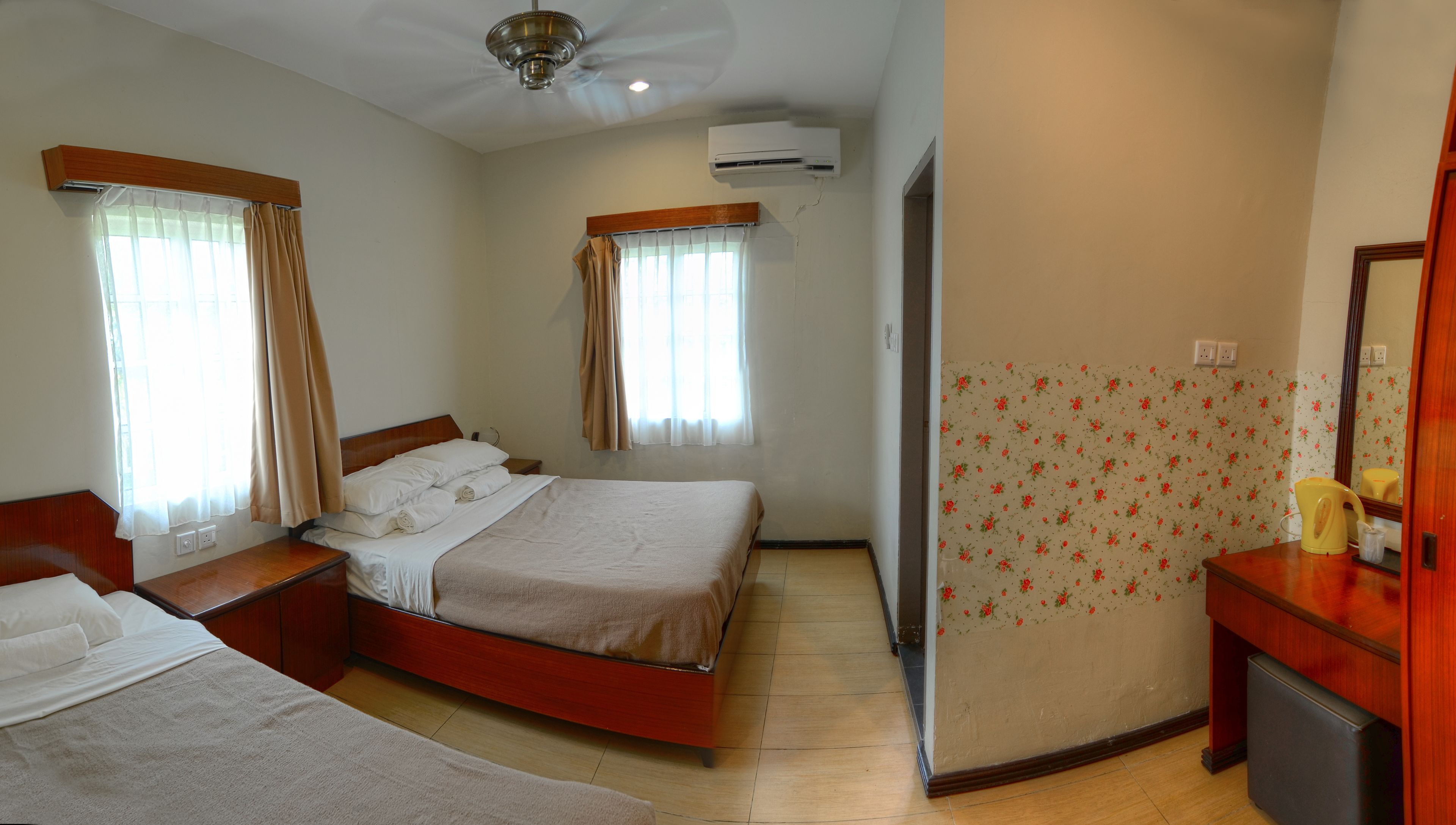 A hotel room with two beds, a ceiling fan, an air conditioning unit, a bathroom with a mirror, and floral wallpaper.
