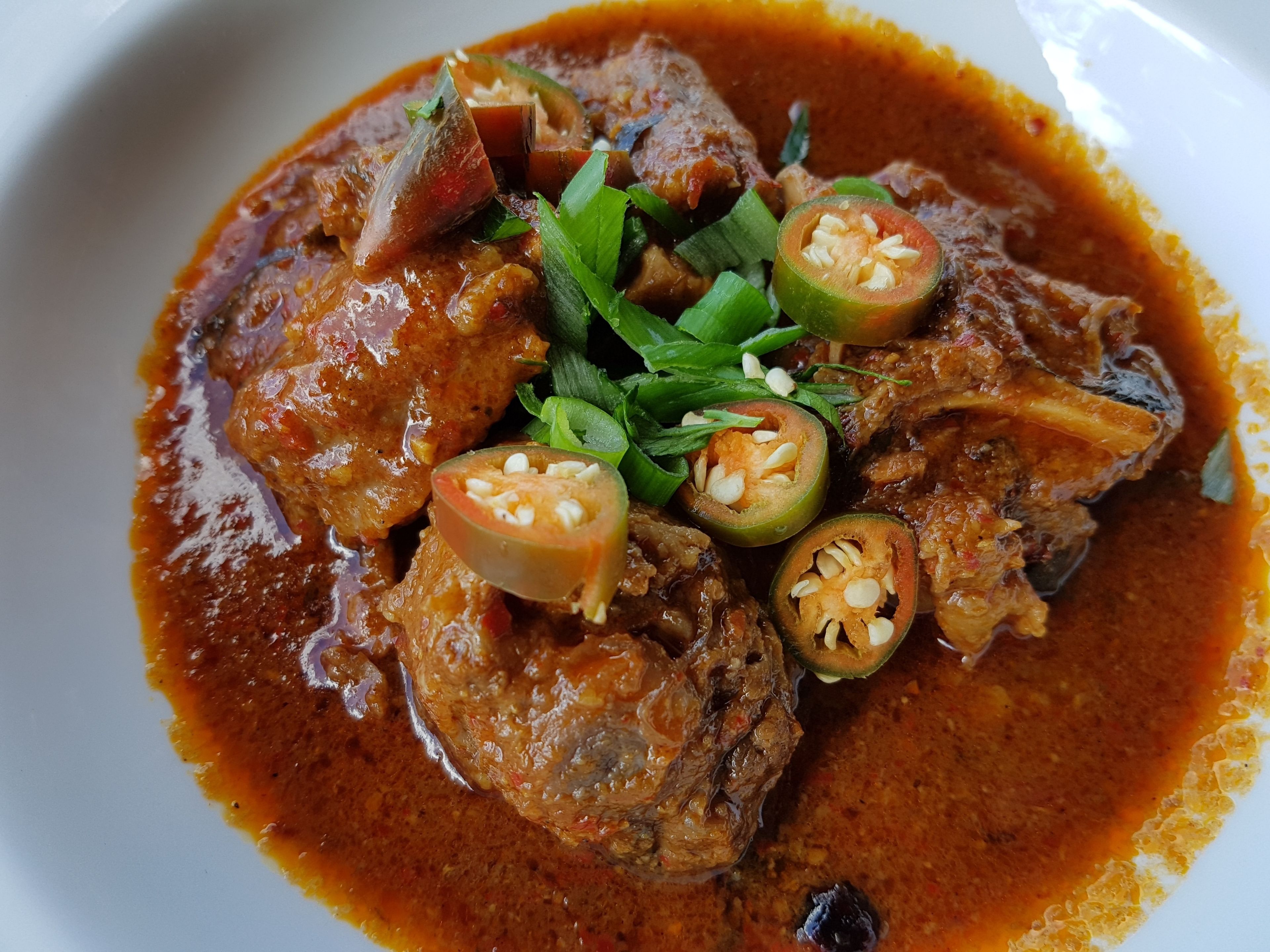 A dish of meat in a rich, reddish sauce, garnished with green herbs and sliced jalapeños.