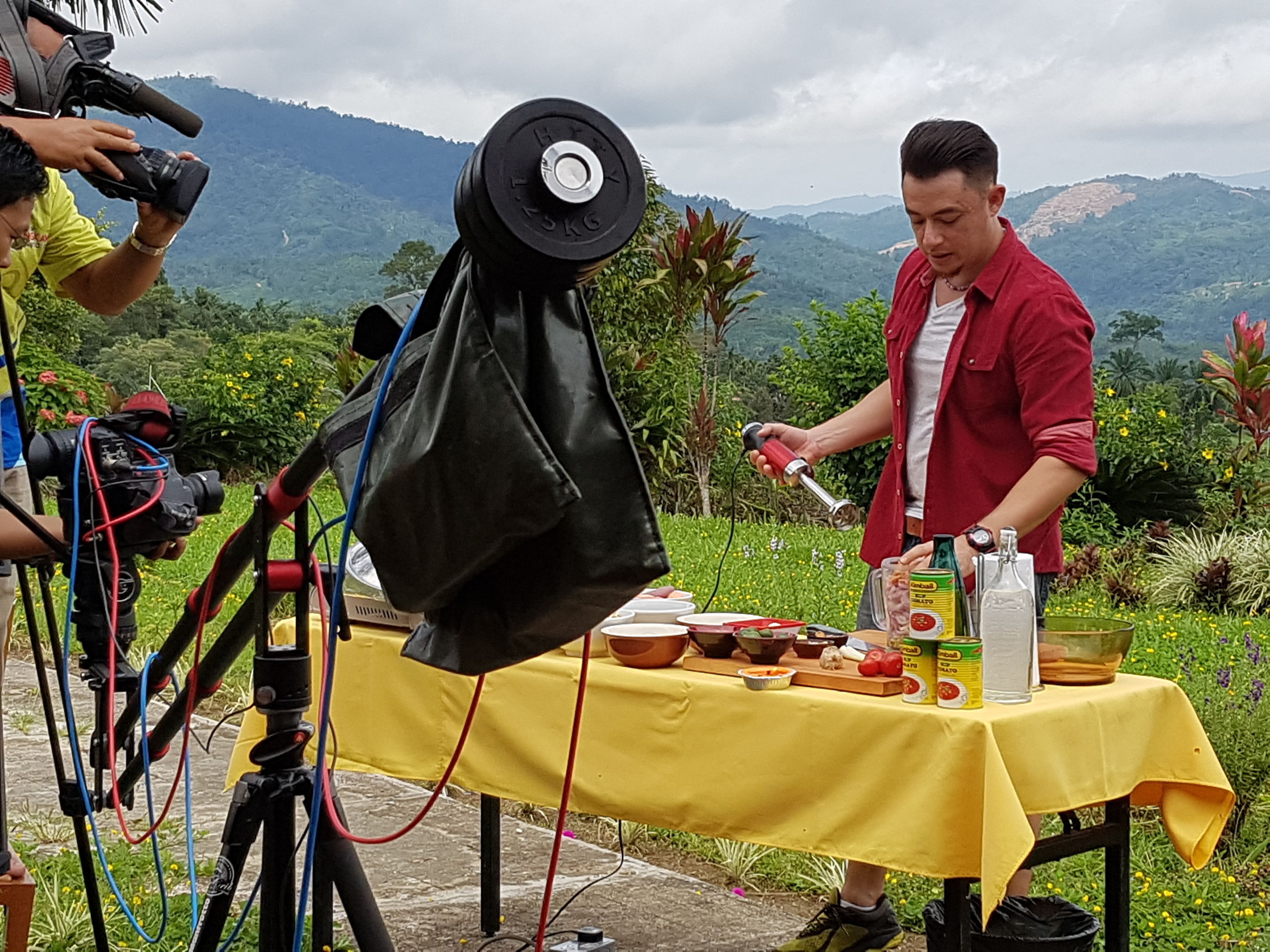 A person cooking outdoors with a table set up, surrounded by greenery and mountains in the background.