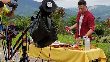 A person cooking outdoors with a table set up, surrounded by greenery and mountains in the background.