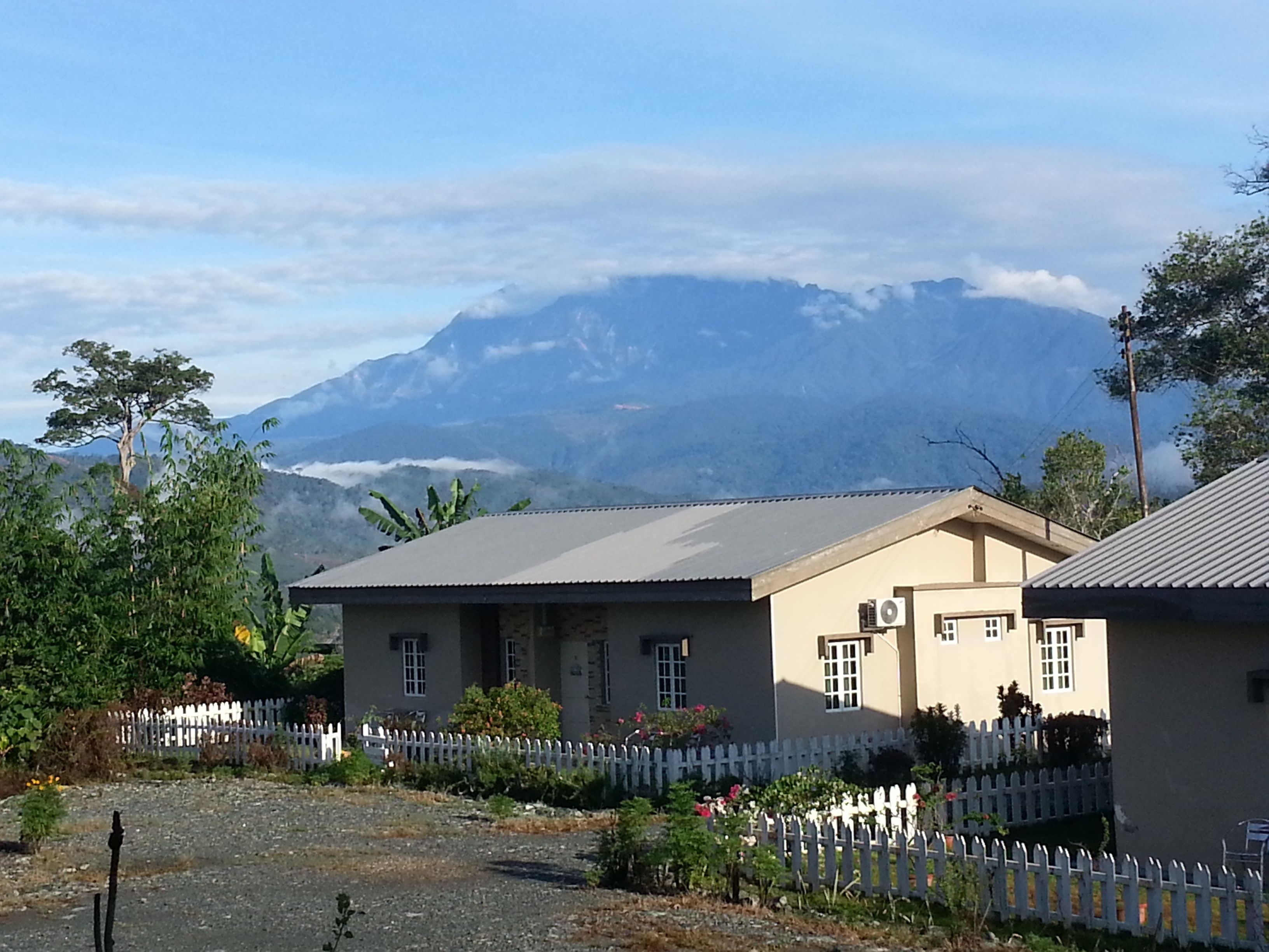 A two-story building with a flat roof, surrounded by a white picket fence, set against a backdrop of mountains and clouds.