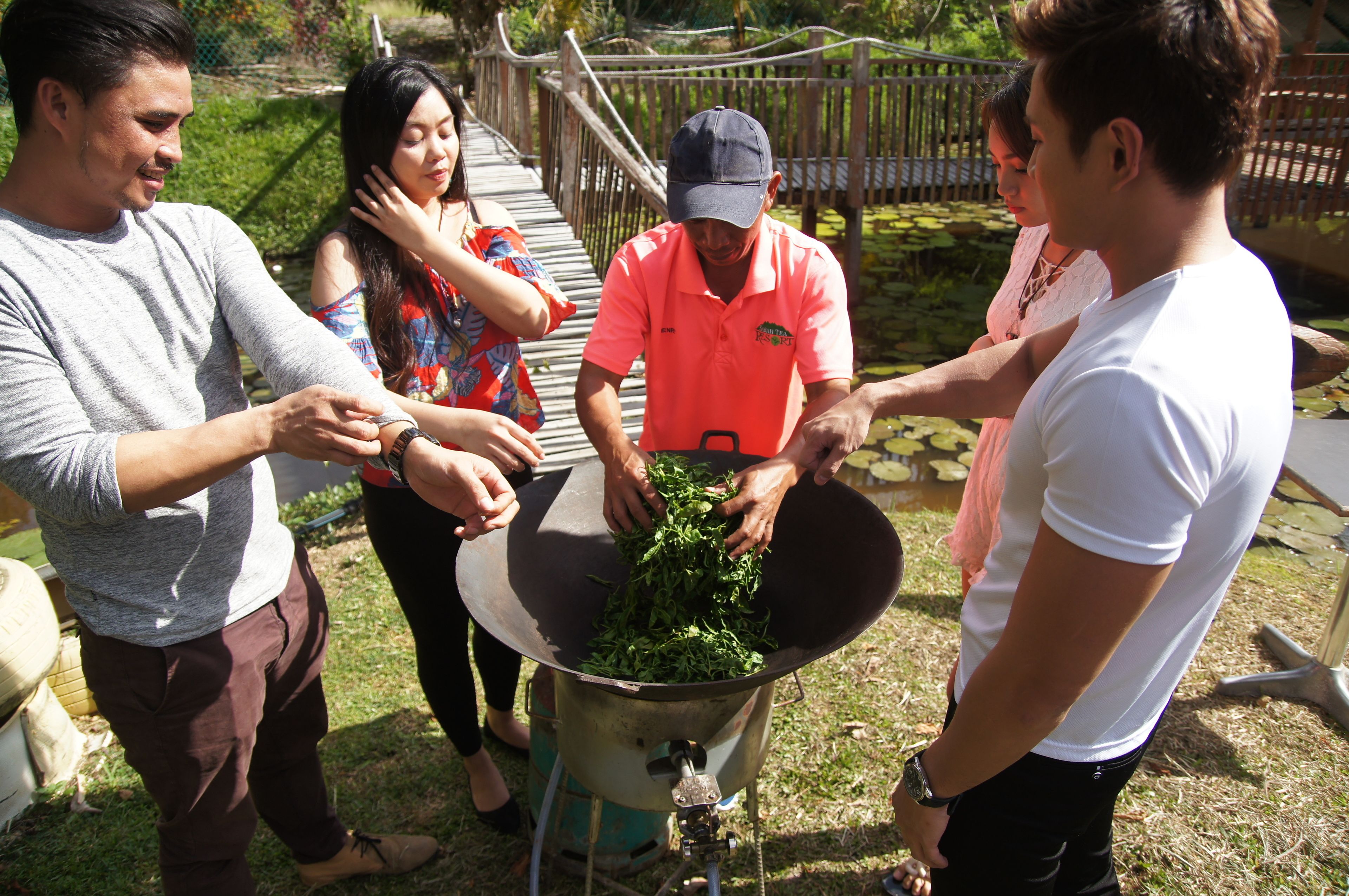 Un grupo de personas alrededor de un recipiente metálico grande lleno de vegetales de hoja, posiblemente preparando comida al aire libre.