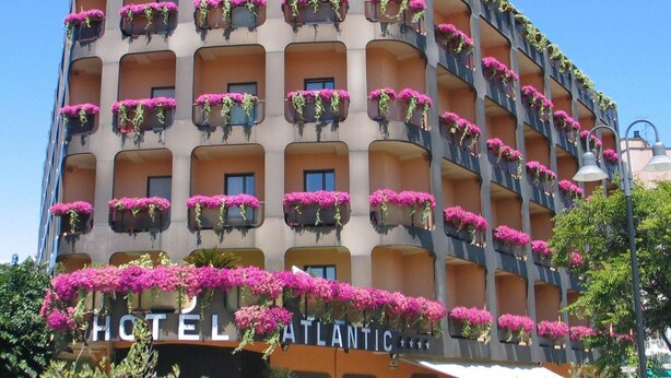 A multi-story hotel with balconies adorned with pink flowers, a visible sign reading "HOTEL ATLANTIC," and a clear blue sky in the background. - Hotel Atlantic (Arona)