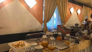 Montaje de bufé con gran variedad de alimentos, frutas y bebidas, en un salón con paredes naranja y ventanas amplias.