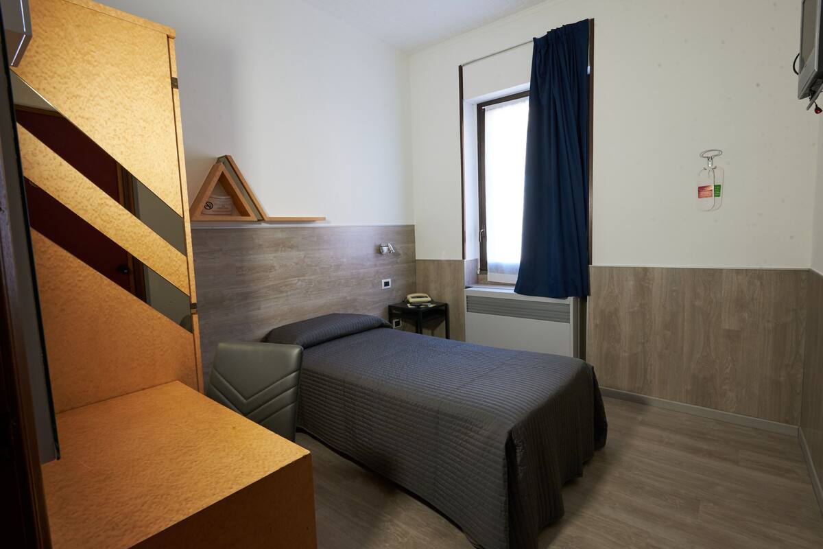 A compact room with a bed, a small bedside table, a chair, a wooden headboard, a window with curtains, and a wall-mounted telephone.