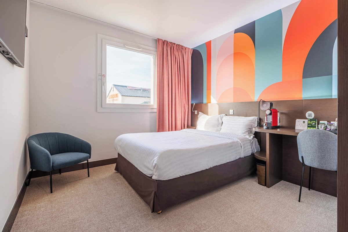 Une chambre d’hôtel équipée d’un lit, d’un bureau, d’une chaise, d’une fenêtre et d’une fresque murale aux couleurs vives.