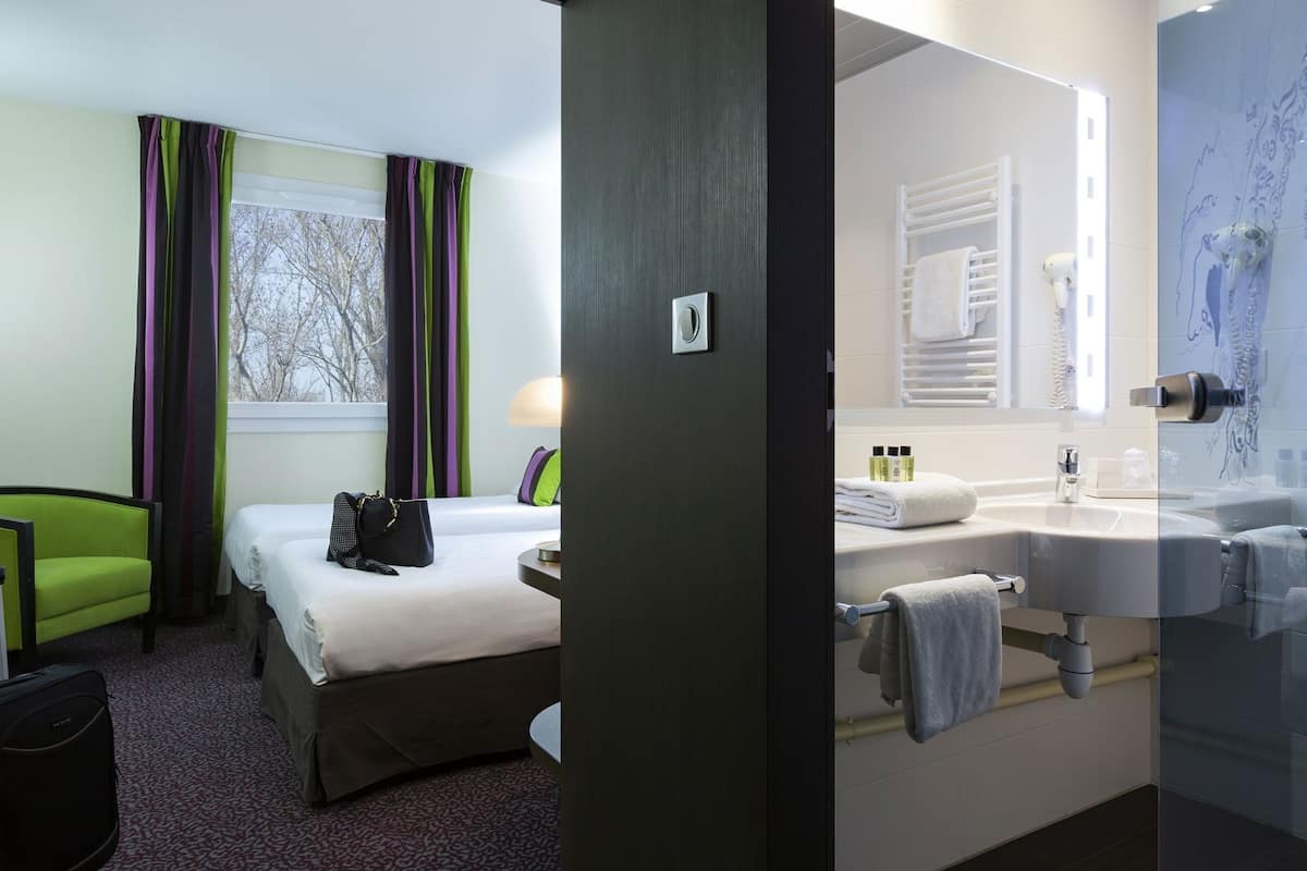 Une chambre d’hôtel comprenant un lit, une chaise, un bureau, une salle de bain avec un lavabo et un miroir, et une fenêtre avec des rideaux.