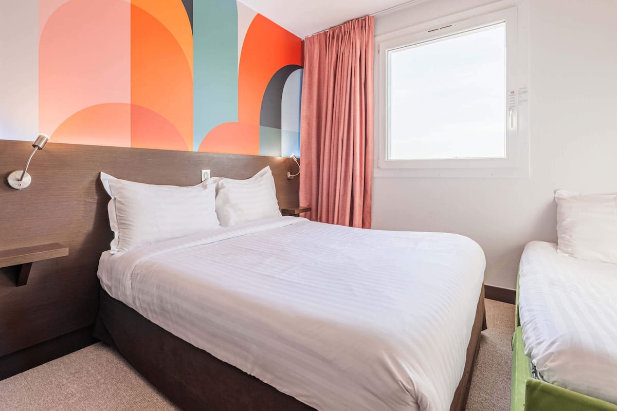 Une chambre d’hôtel avec un grand lit, une table de chevet, une lampe fixée au mur et une fenêtre avec des rideaux.