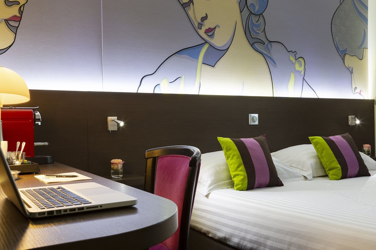 Une chambre d’hôtel avec un lit, un bureau avec un ordinateur portable, une chaise et un mur orné d’une fresque.