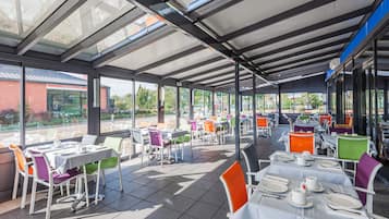 An outdoor dining area with tables and chairs, covered by a glass roof, and a view of the street and greenery.