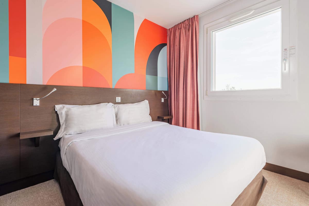 Une chambre d’hôtel avec un grand lit, une tête de lit en bois, une fresque murale abstraite aux couleurs vives et une fenêtre avec des rideaux.