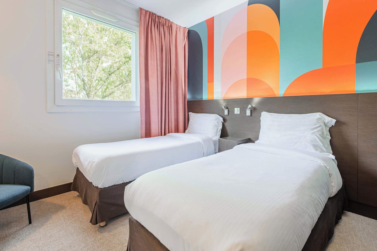 Une chambre d’hôtel avec deux lits simples, une fenêtre donnant sur la verdure et un motif mural abstrait aux couleurs vives.