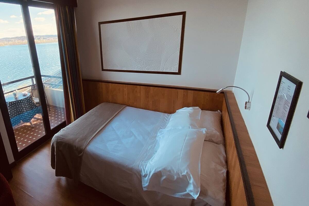 Habitación de hotel con cama, ventana con vista al mar, cabecera de madera y un cuadro enmarcado en la pared.