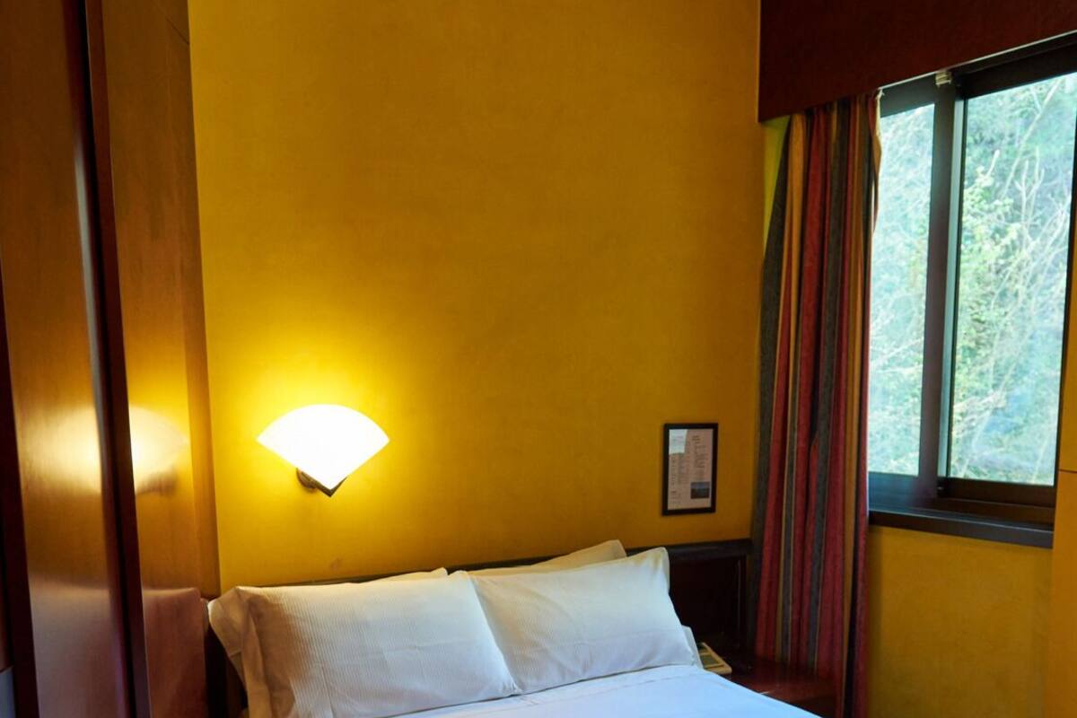 Habitación de hotel con cama, luz empotrada, ventana con cortinas e imagen enmarcada en la pared.