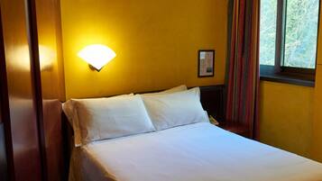 A hotel room with a bed, a wall-mounted light, a window with curtains, and a framed picture on the wall.