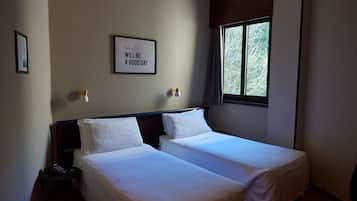 A room with two single beds, a framed motivational quote on the wall, a small table with a phone, and a window with a view of trees.