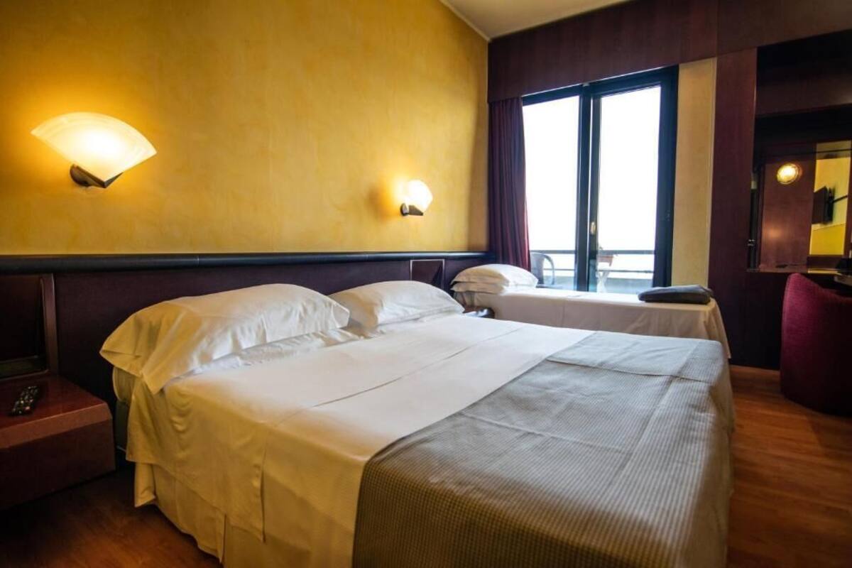 Una habitación de hotel con una cama grande, una mesita de noche, una silla y un ventanal con cortinas.