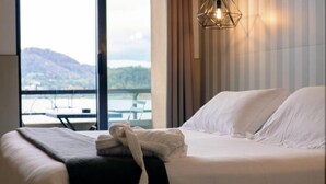 A hotel room with a bed, a window with a view of a body of water and mountains, and a hanging light fixture. - Hotel Concorde (Arona)