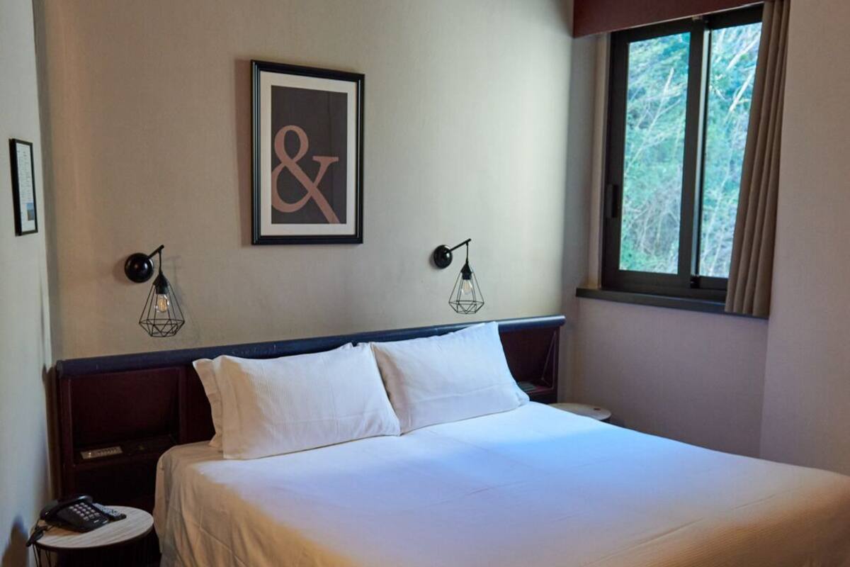 Una cama bien tendida en una habitación de hotel con un ventanal grande, una obra de arte enmarcada en la pared y dos lámparas fijadas a la pared.