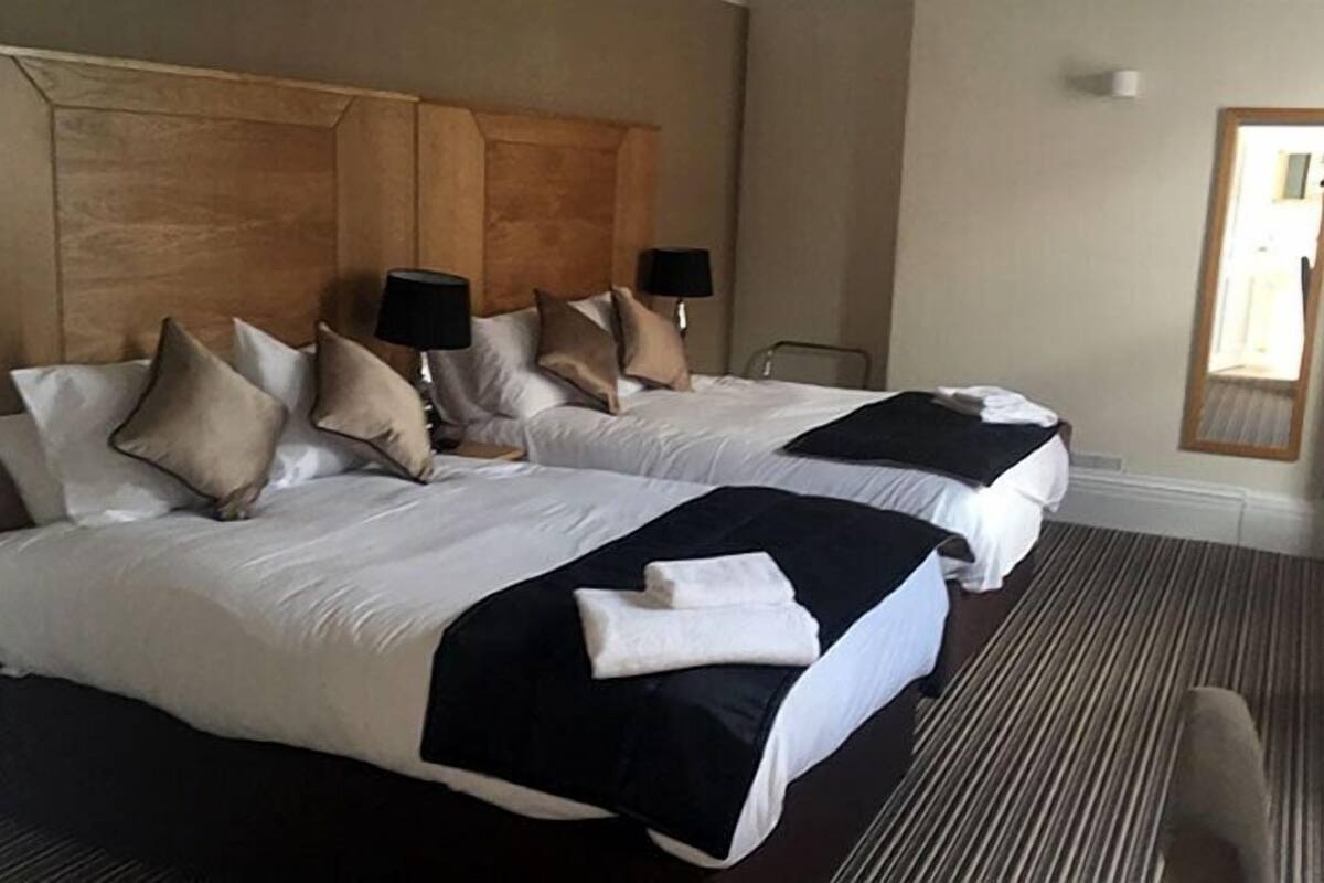 A hotel room with two beds, each with a headboard, pillows, and lamps. There is a carpeted floor and a door visible.
