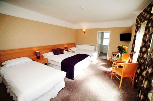 A hotel room with two single beds, a wooden headboard, a small table with a chair, a TV mounted on the wall, and a window with curtains.
