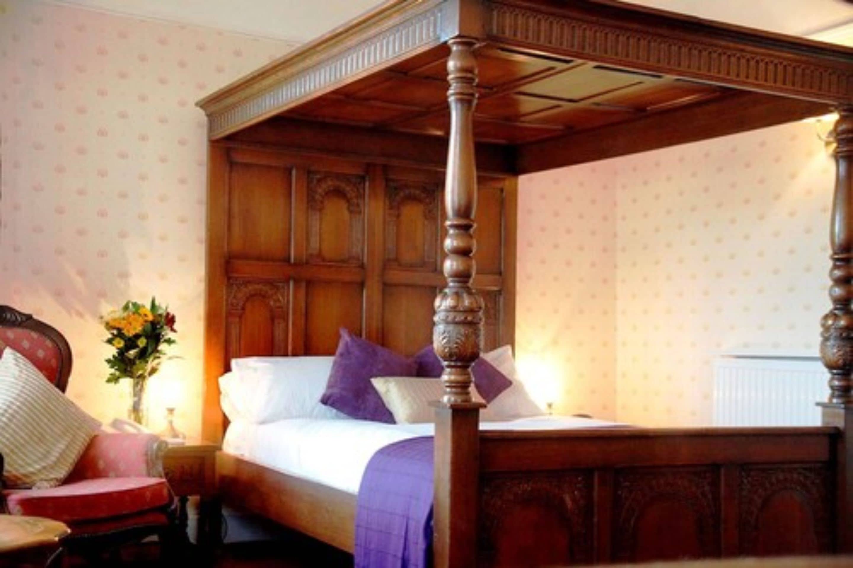 Una cama con dosel y tablero de madera, una mesita de noche con un florero y una silla.