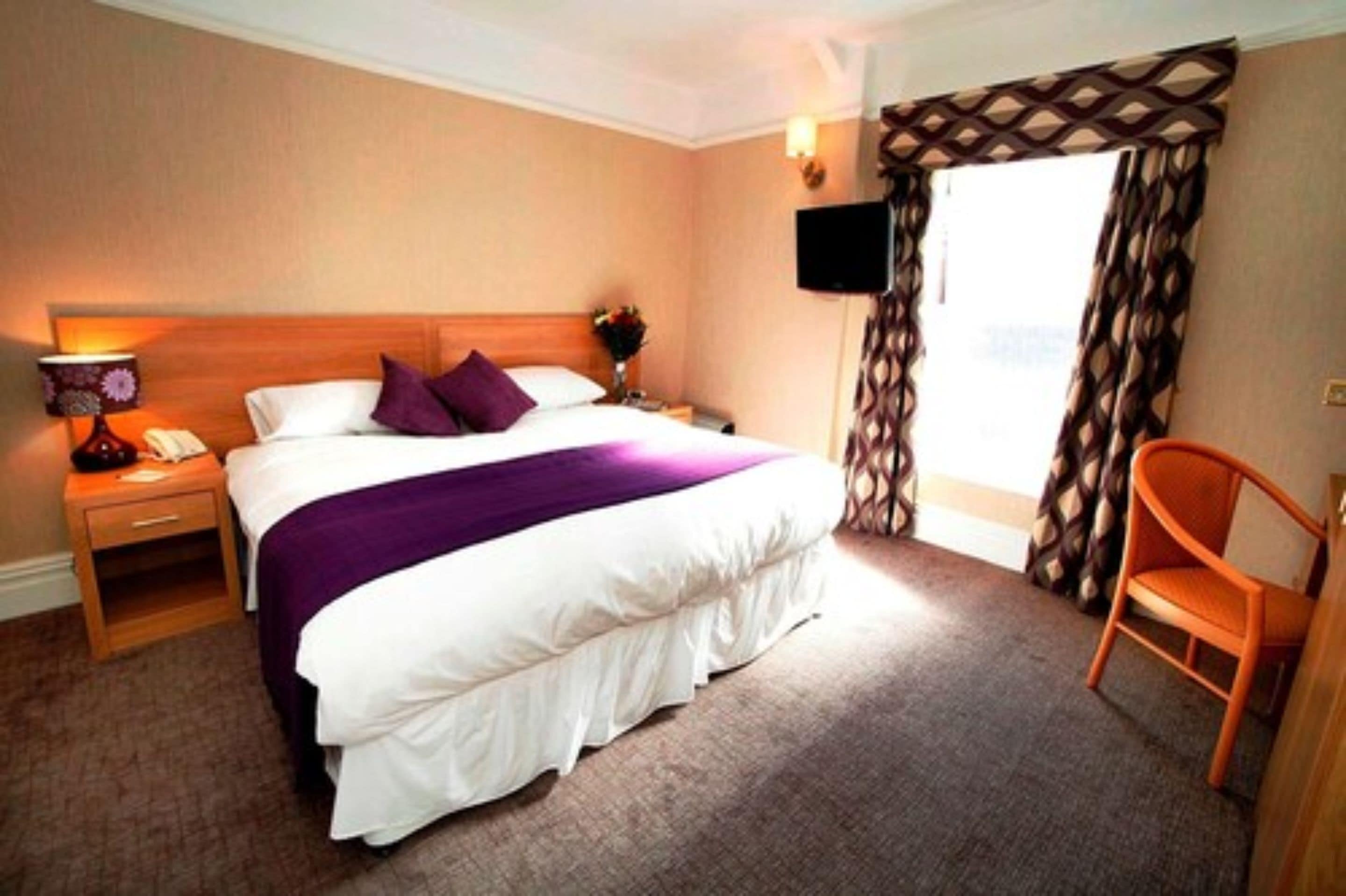 A hotel room with a large bed, a nightstand with a lamp, a chair, and a TV mounted on the wall.
