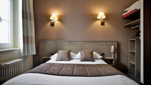 A hotel room with a large bed, two bedside lamps, a wooden headboard, a bedside telephone, and a built-in shelf with clothes. - Brit Hotel Le Surcouf (Saint-Malo)