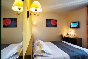 Quarto de hotel com duas camas, uma escrivaninha, uma televisão e duas luminárias.
